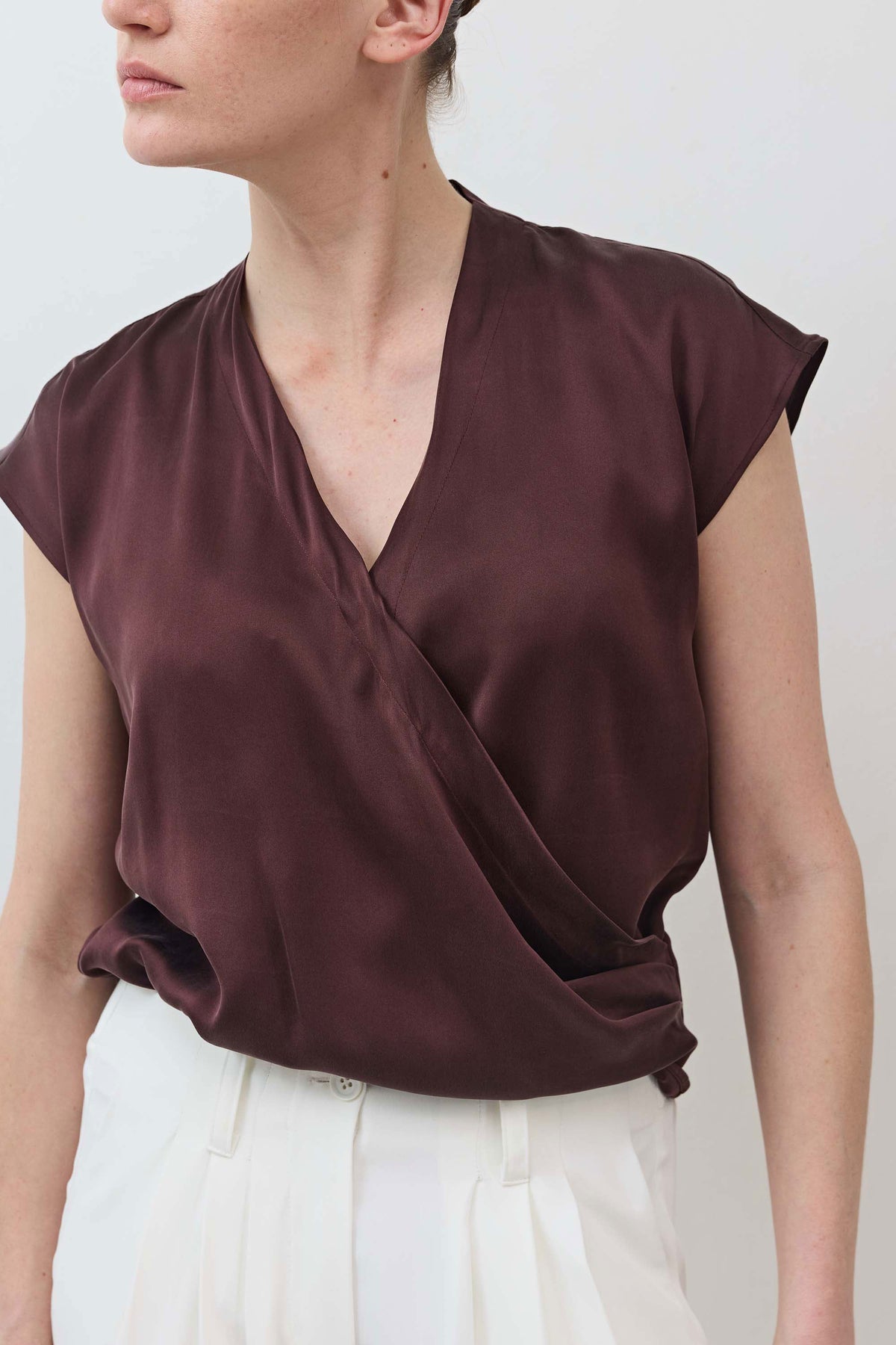 Detail view of fudge brown wrap silk top showing V-neck and diagonal drape over white trousers.